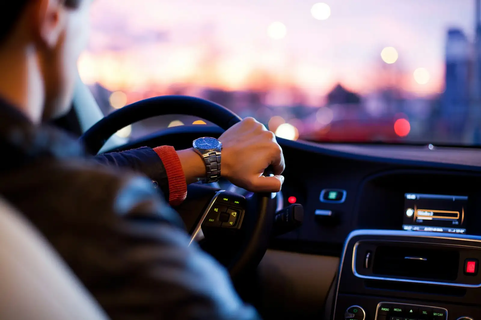 Vista desde el asiento del conductor, manos sobre el volante, al atardecer con las luces de la ciudad desenfocadas al fondo.